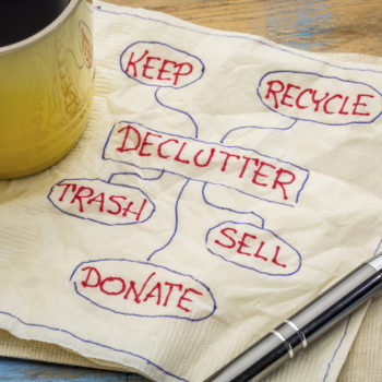 A square white paper napkin with a yellow coffee cup resting on one side of it and a pen resting on the other side. Written on the napkin in pen is the word "Declutter" in a box with the following words surrounding it: Keep, Recycle, Trash, Sell, Donate. There is a circle around each word and a line drawn from the Declutter box to each circle.