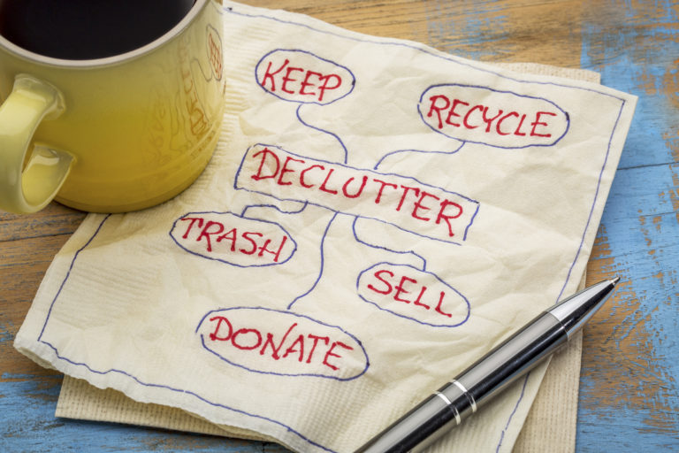 A square white paper napkin with a yellow coffee cup resting on one side of it and a pen resting on the other side. Written on the napkin in pen is the word "Declutter" in a box with the following words surrounding it: Keep, Recycle, Trash, Sell, Donate. There is a circle around each word and a line drawn from the "Declutter" box to each circle.