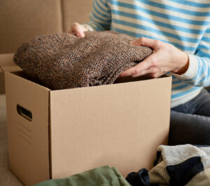 Person wearing a blue-and-white striped shirt folds a brown knitted garment and places it into a cardboard box for packing move.